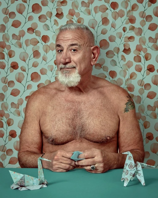 Mature man with white beard folding origami at a table. He has a slight smile and is shirtless, set against a leafy wallpaper.